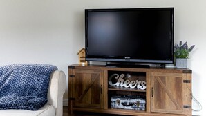 Un salón con una televisión sobre un mueble de madera, un sofá con una manta azul y una pequeña decoración con forma de casa.