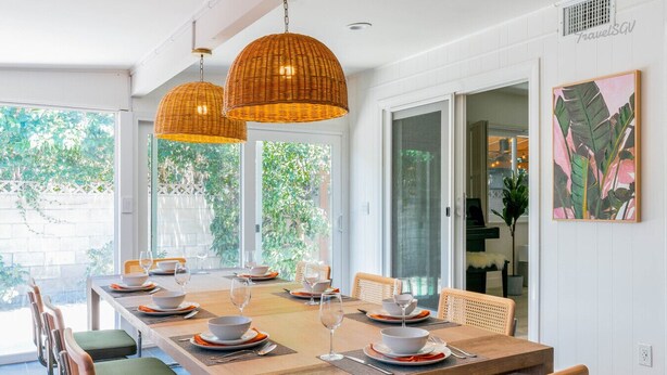 A dining area with a long wooden table set for six, surrounded by wicker chairs, under two large wicker pendant lights.