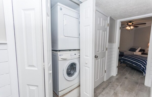 A laundry room with a front-loading washer and dryer, a door leading to a bedroom with a bed and a ceiling fan.