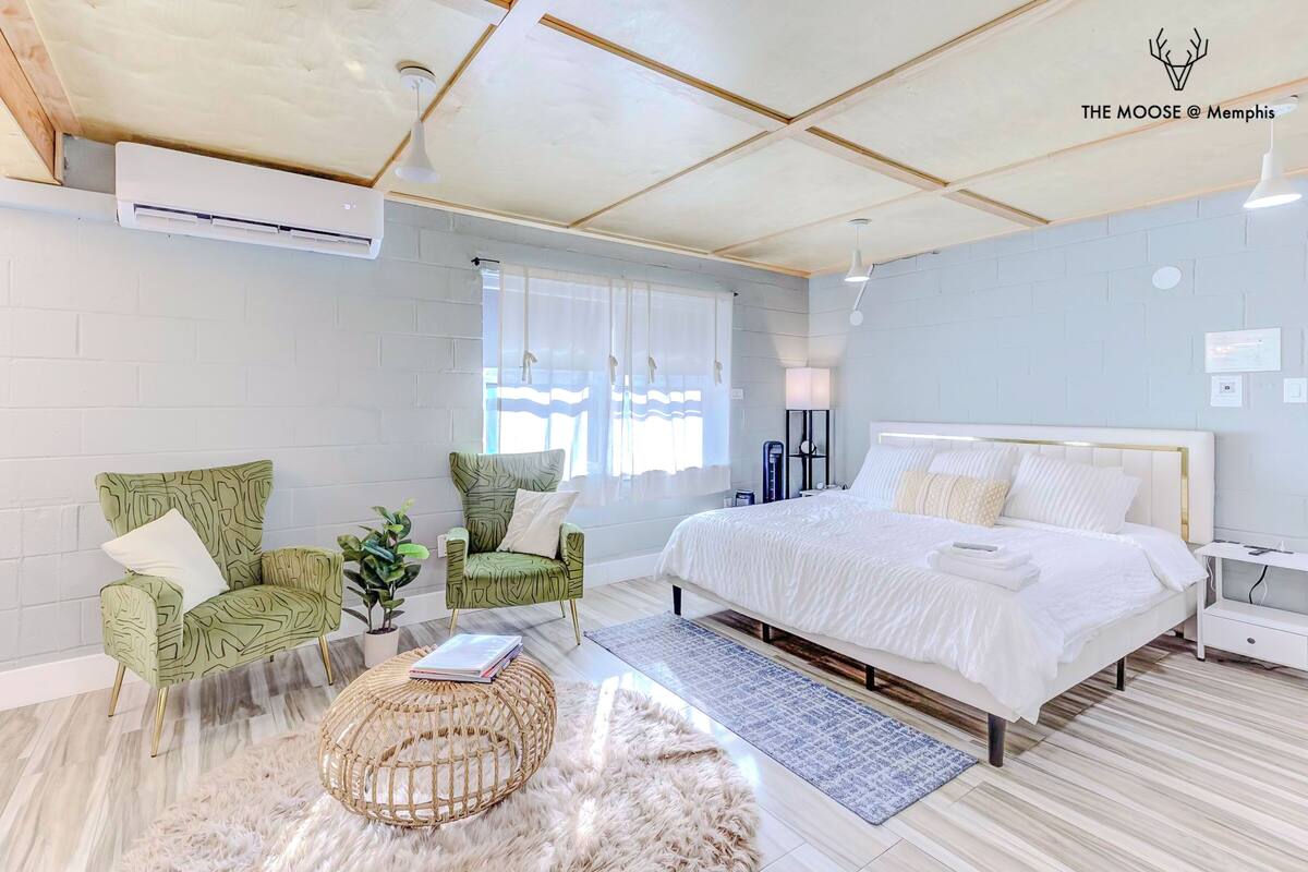 Un dormitorio moderno con una cama grande, dos sillones de estampado verde, una mesita de noche blanca y una alfombra mullida.