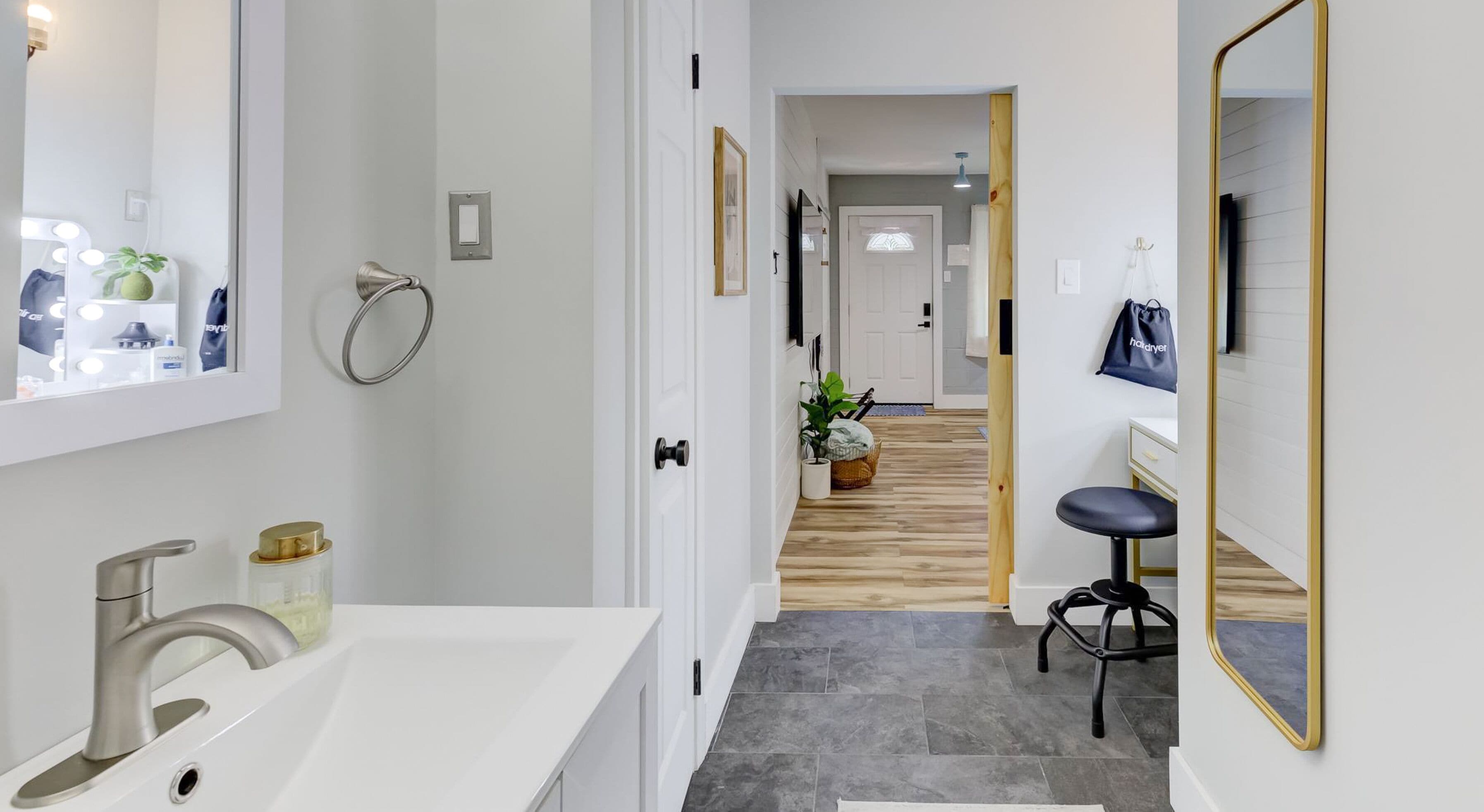 A modern bathroom with a white sink, a mirror, and a wooden floor leading to a hallway.