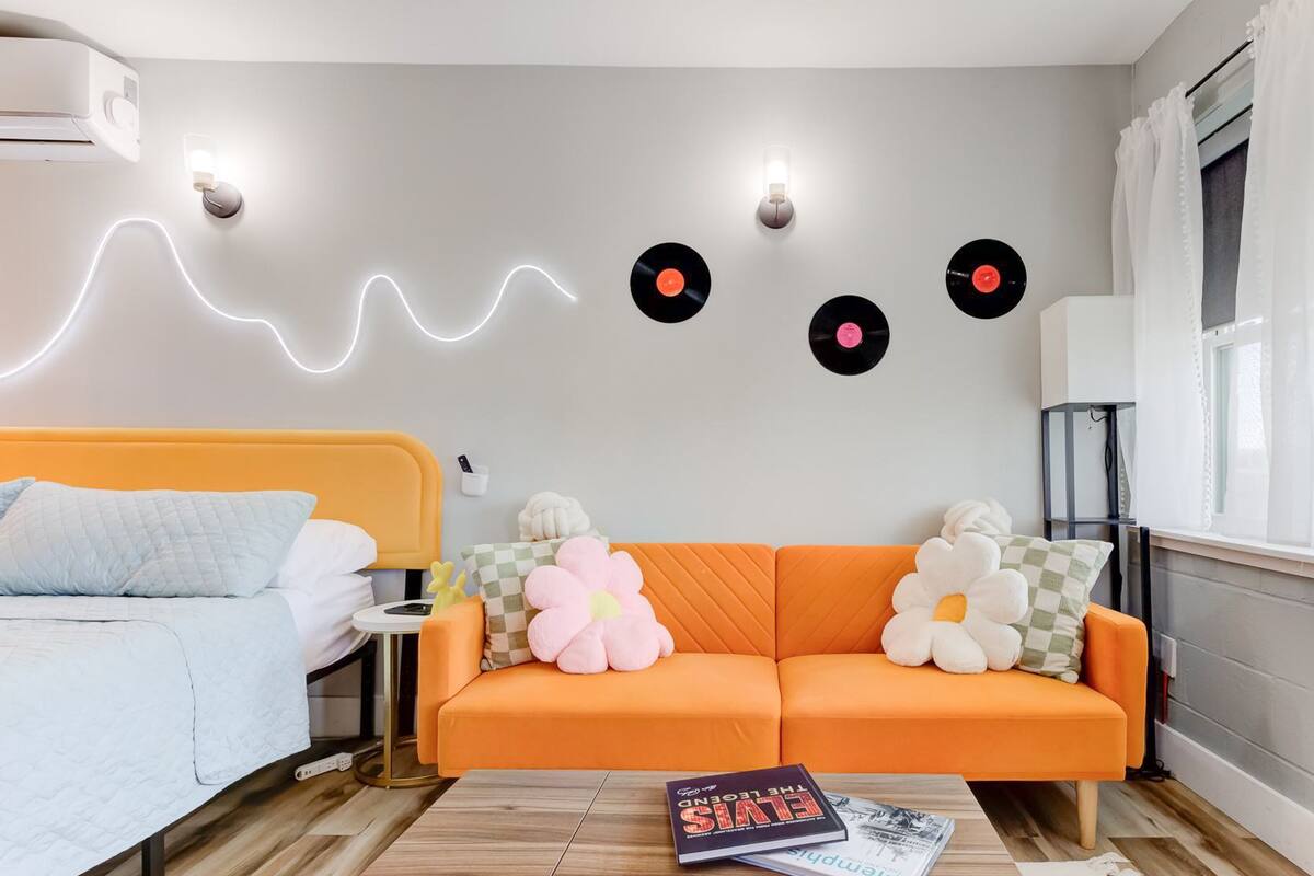 Un dormitorio moderno con cama, sofá naranja, una mesa de centro y adornos de pared que incluyen discos de vinilo y una tira de luces.