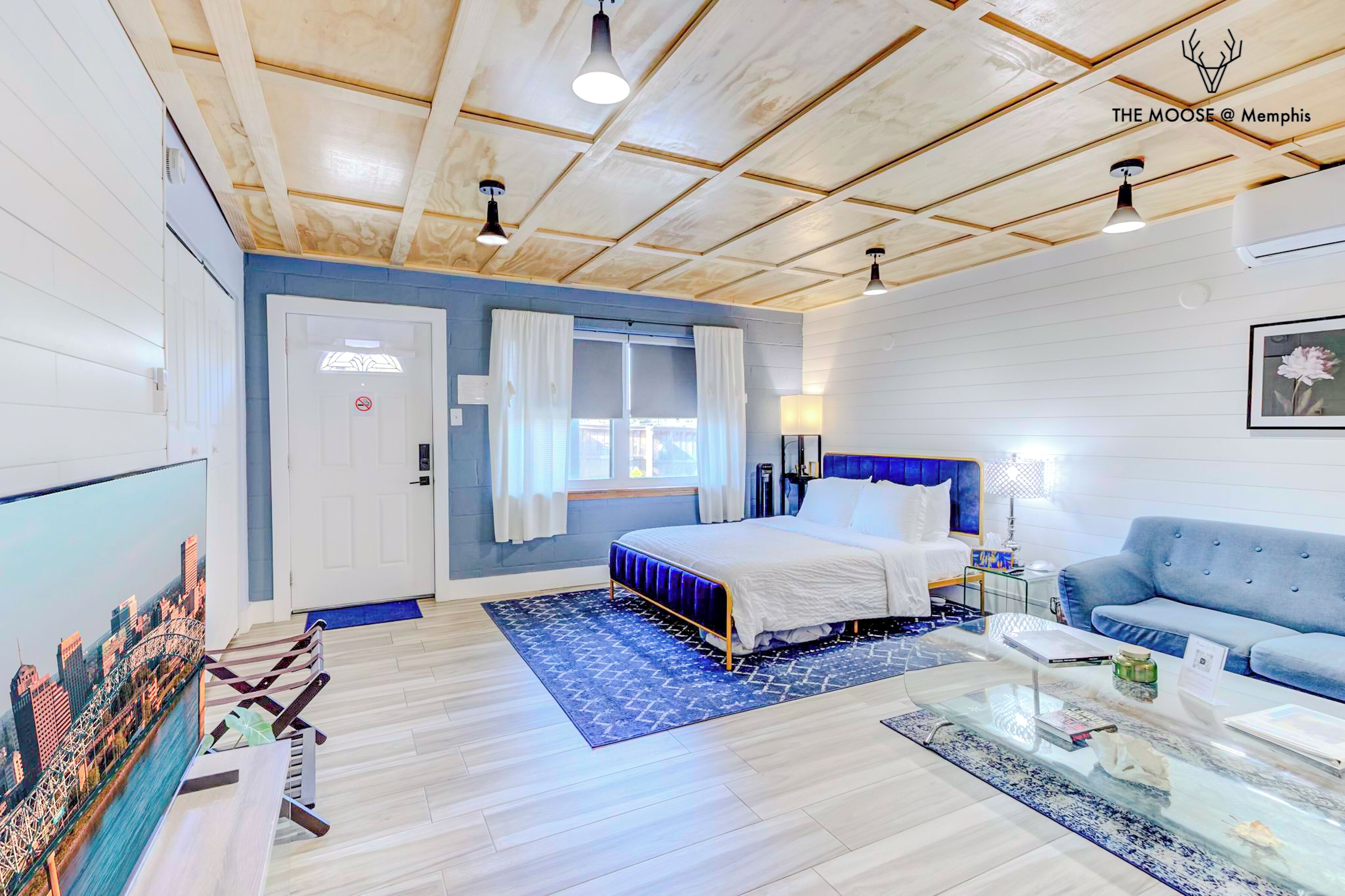 A modern hotel room with a large bed, a blue sofa, a glass coffee table, and a cityscape view on the TV.