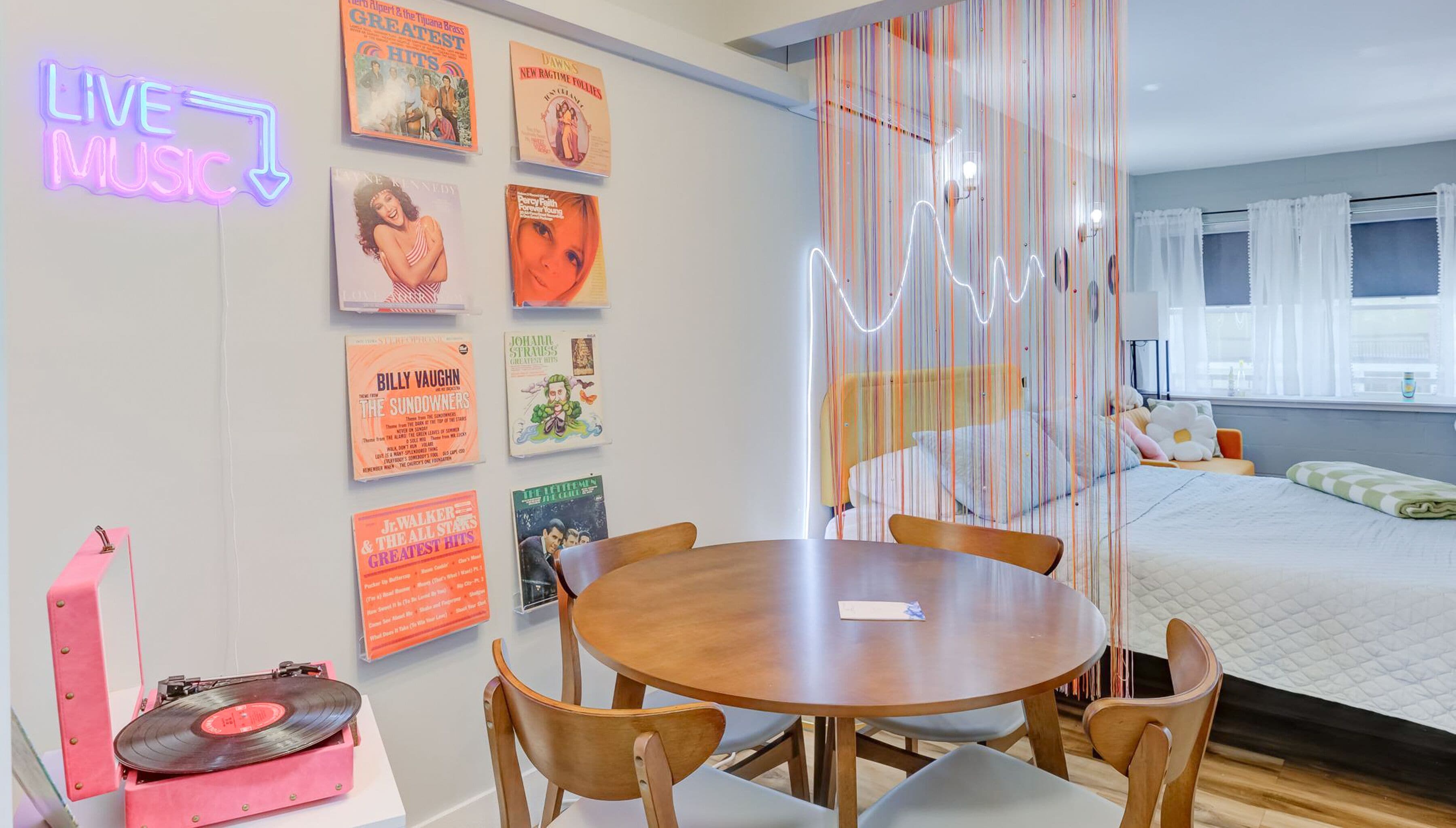 A modern hotel room with a round wooden table, chairs, a pink record player, and a neon sign indicating a live music area.