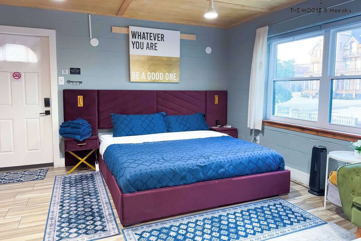 Un dormitorio con cama, mesita de noche, silla, ventana y un cartel en la pared.