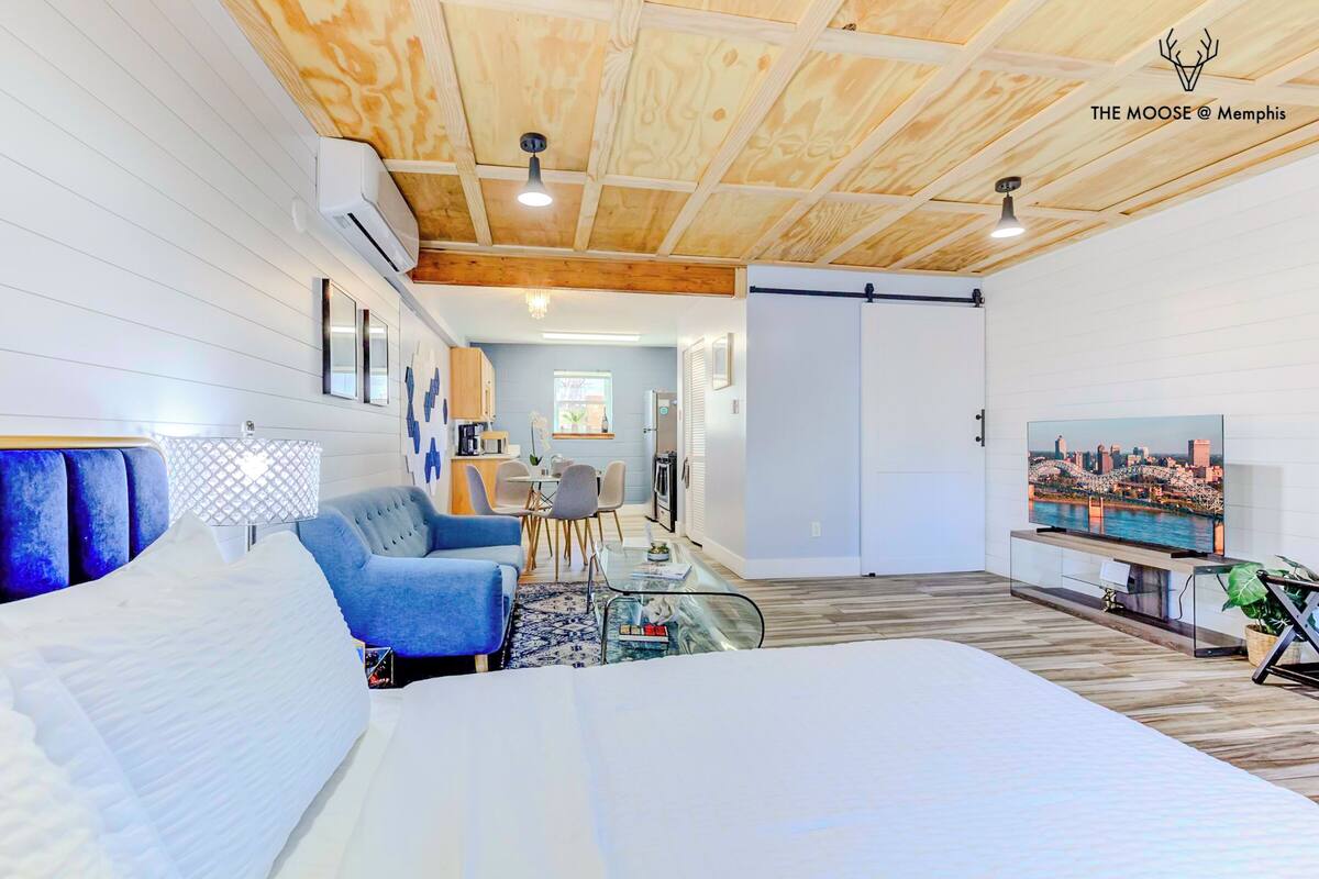 Una sala moderna con una cama, un sillón azul y un televisor mostrando un paisaje urbano.