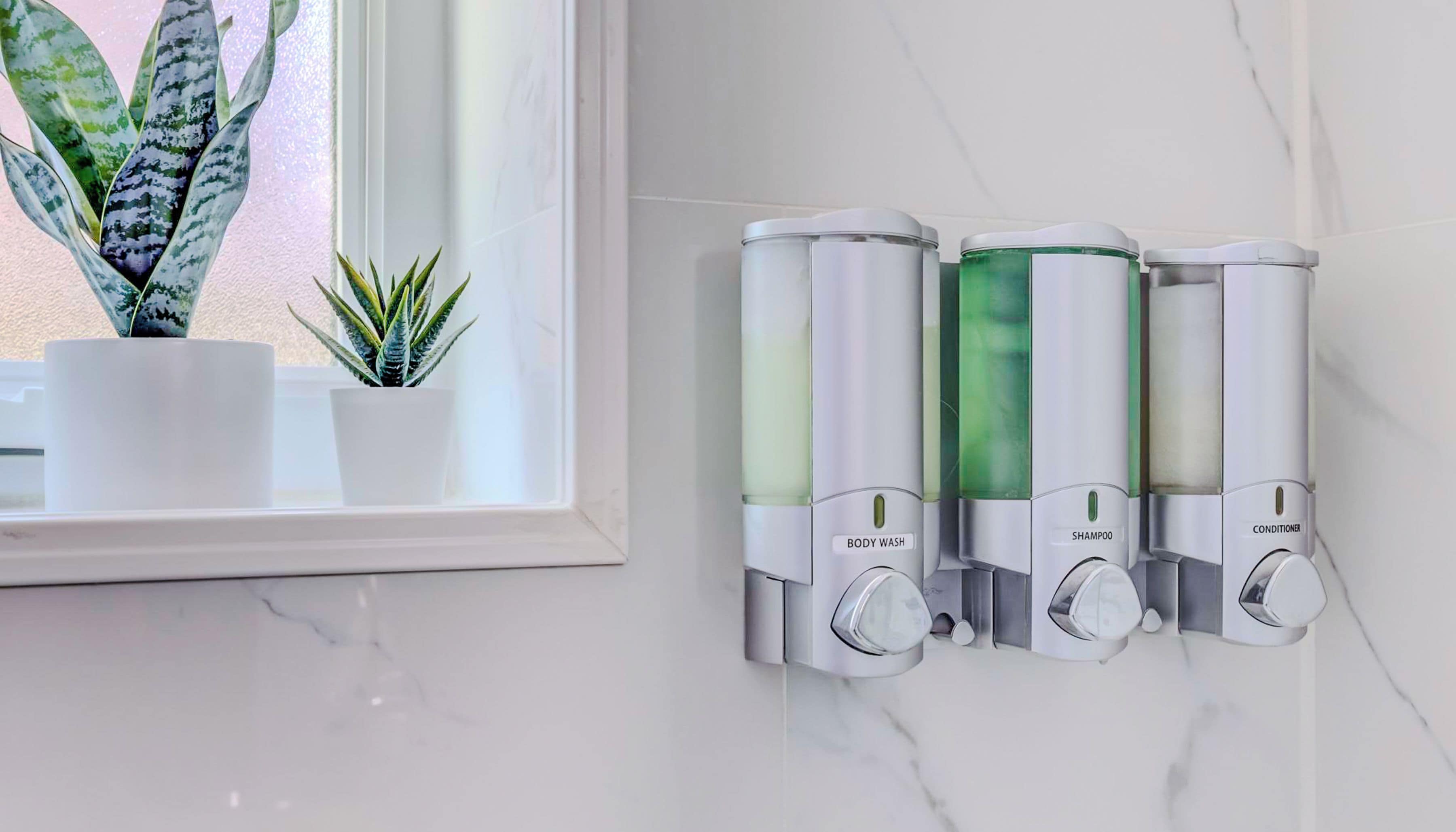 A bathroom with a marble countertop, a potted plant, and three wall-mounted dispensers labeled body wash, shampoo, and conditioner.