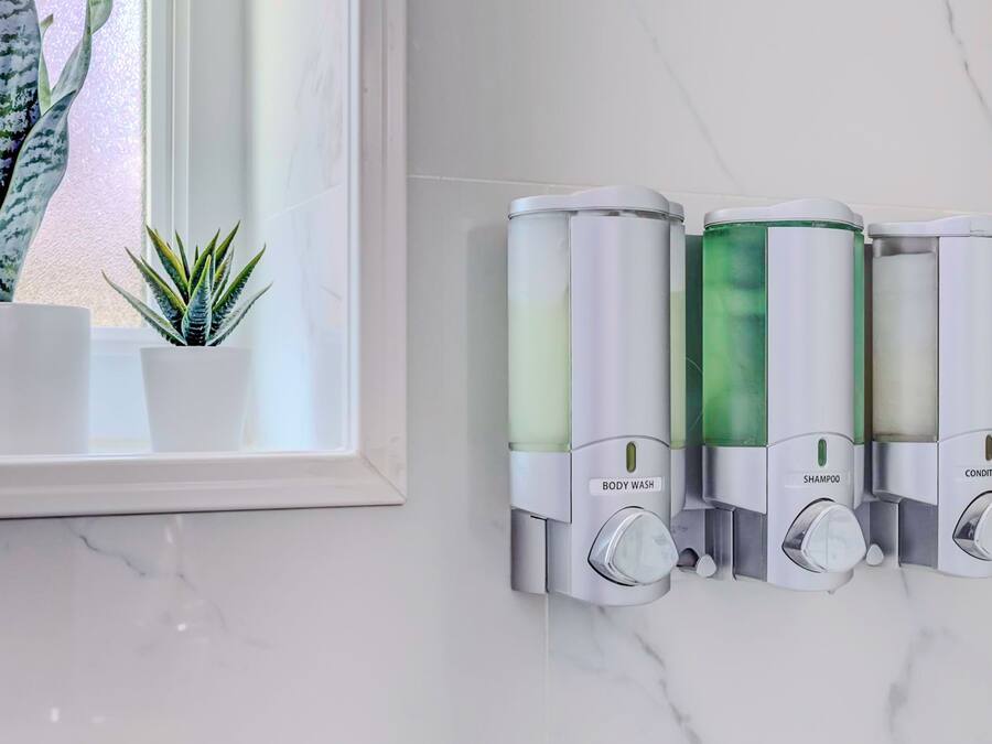 A bathroom with a marble countertop, a potted plant, and three wall-mounted dispensers labeled body wash, shampoo, and conditioner.