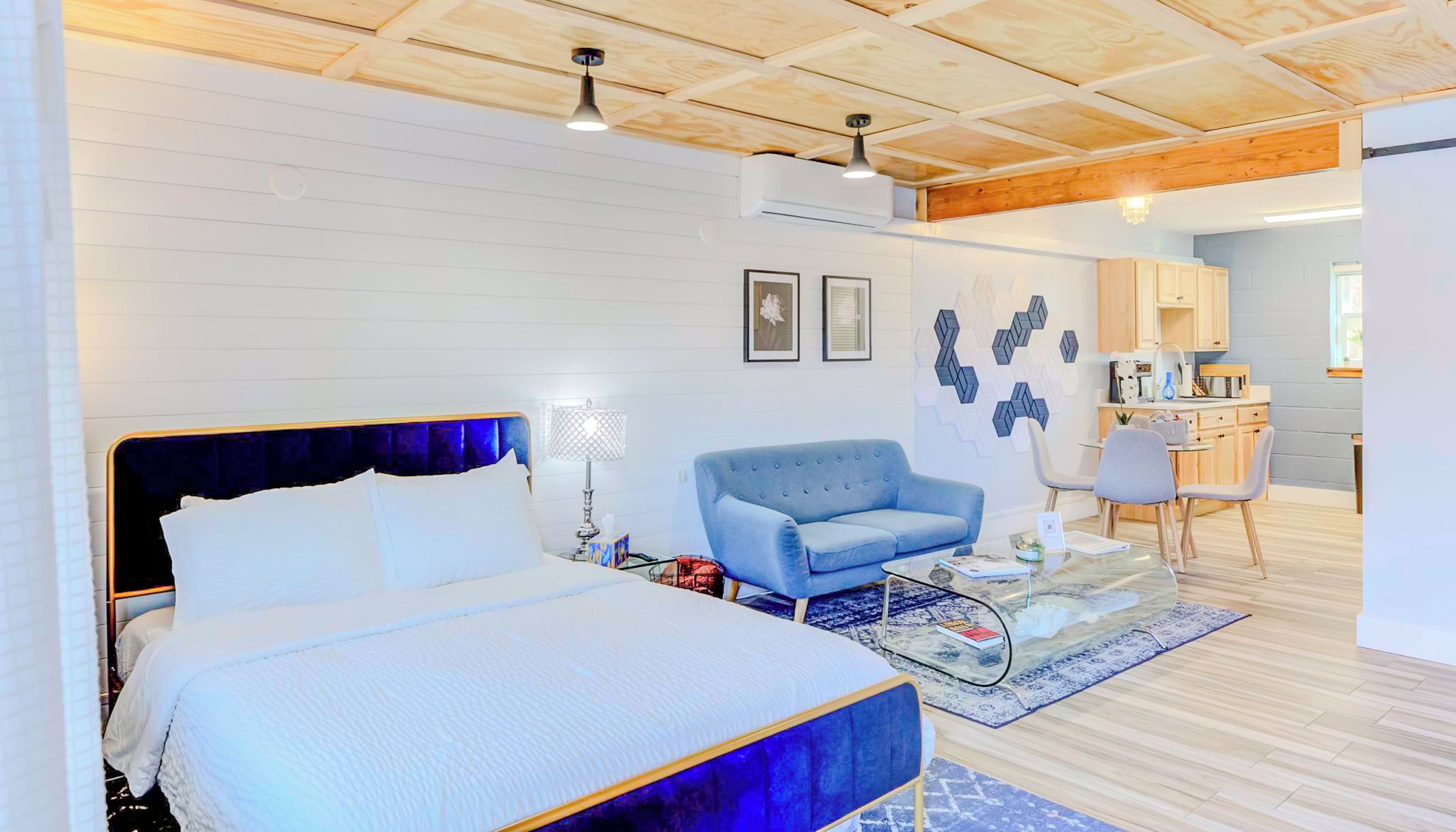 A modern hotel room with a large bed, a blue armchair, a small table, and a kitchenette in the background.