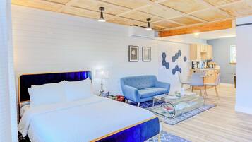 A modern hotel room with a large bed, a blue armchair, a small table, and a kitchenette in the background.