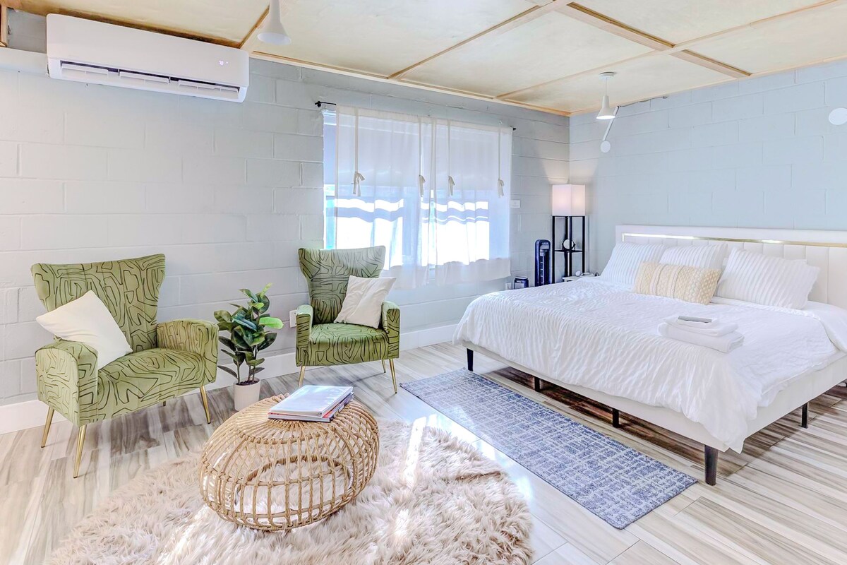 Un dormitorio moderno con una cama grande, dos sillones de estampado verde, una mesita de noche blanca y una alfombra mullida.