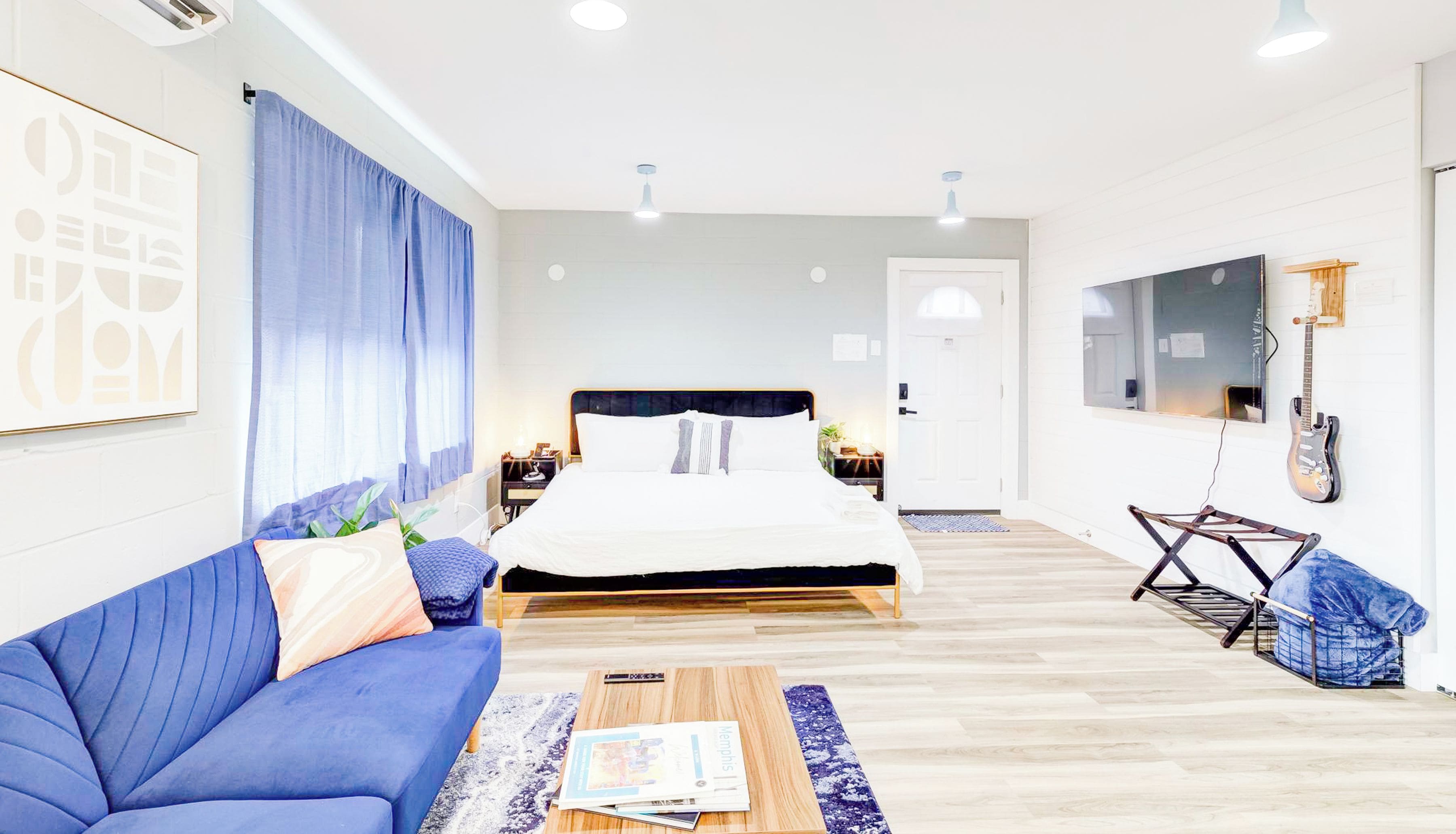 Una habitación de hotel moderna con una cama, un sofá azul, una mesa de centro de madera, un televisor y una guitarra.