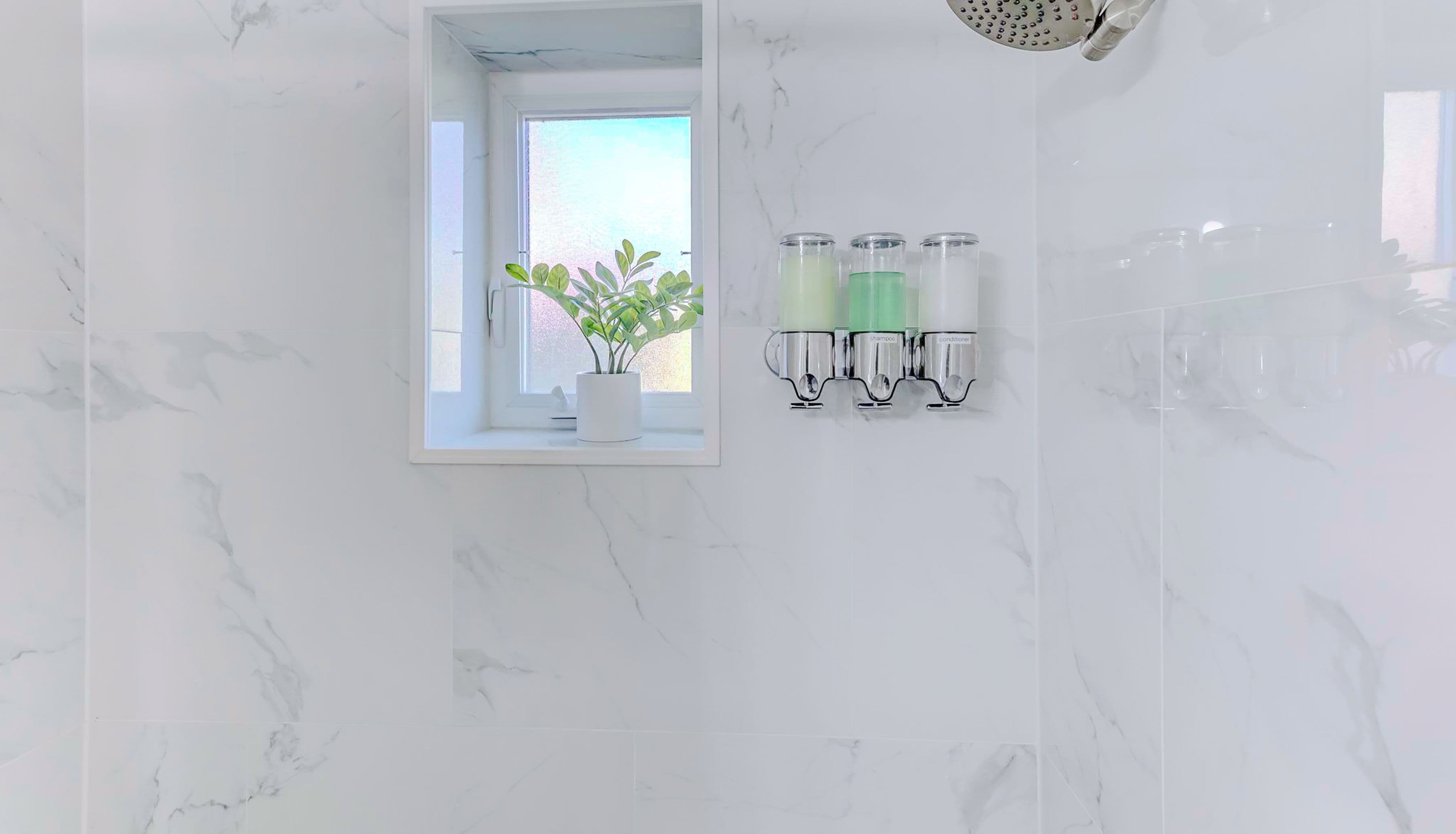 Un baño con paredes de mármol blanco, una planta en maceta, una ducha y tres recipientes de vidrio en la pared.