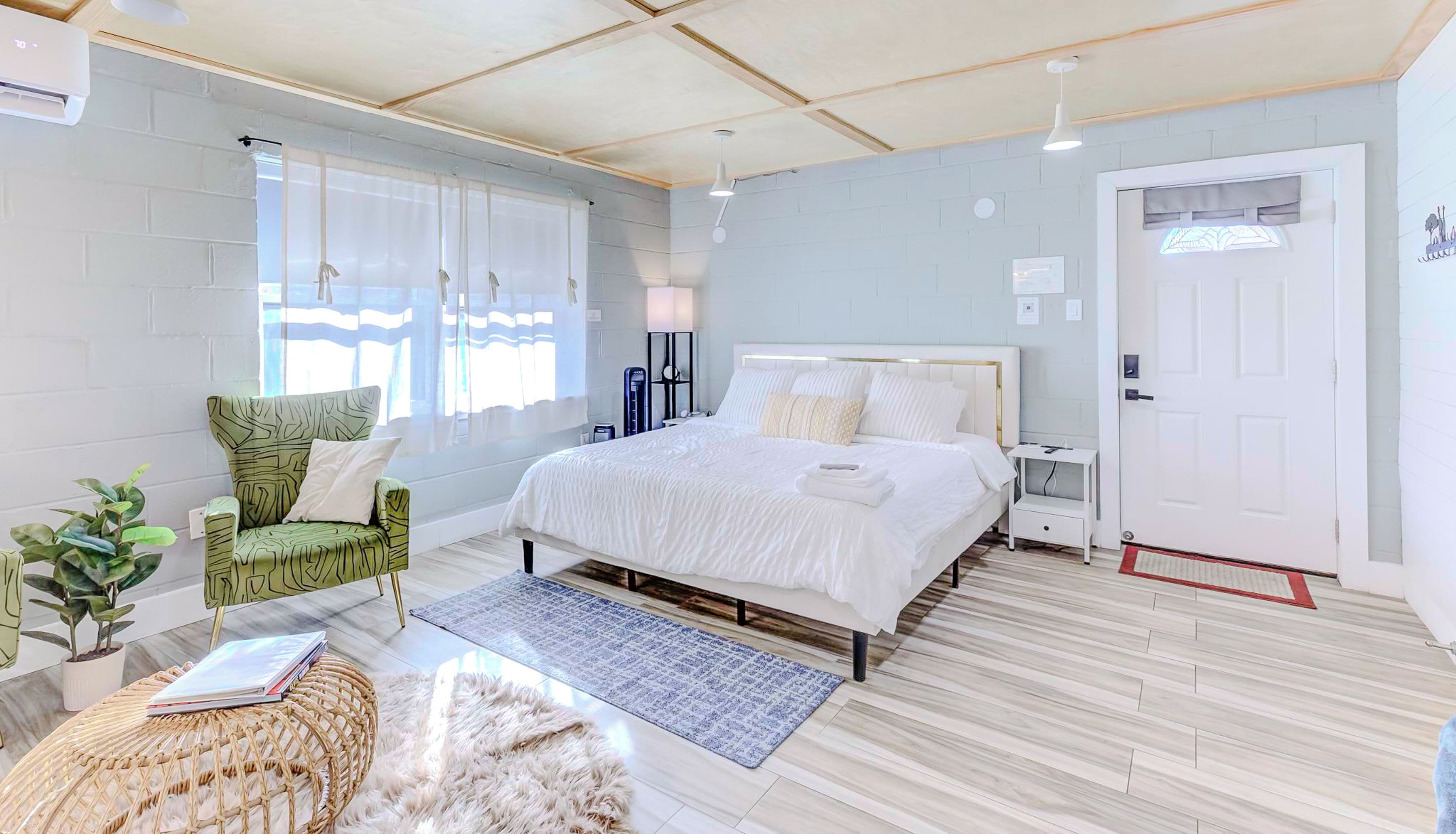 A modern bedroom with a large bed, a green patterned chair, a small bedside table, and a potted plant.