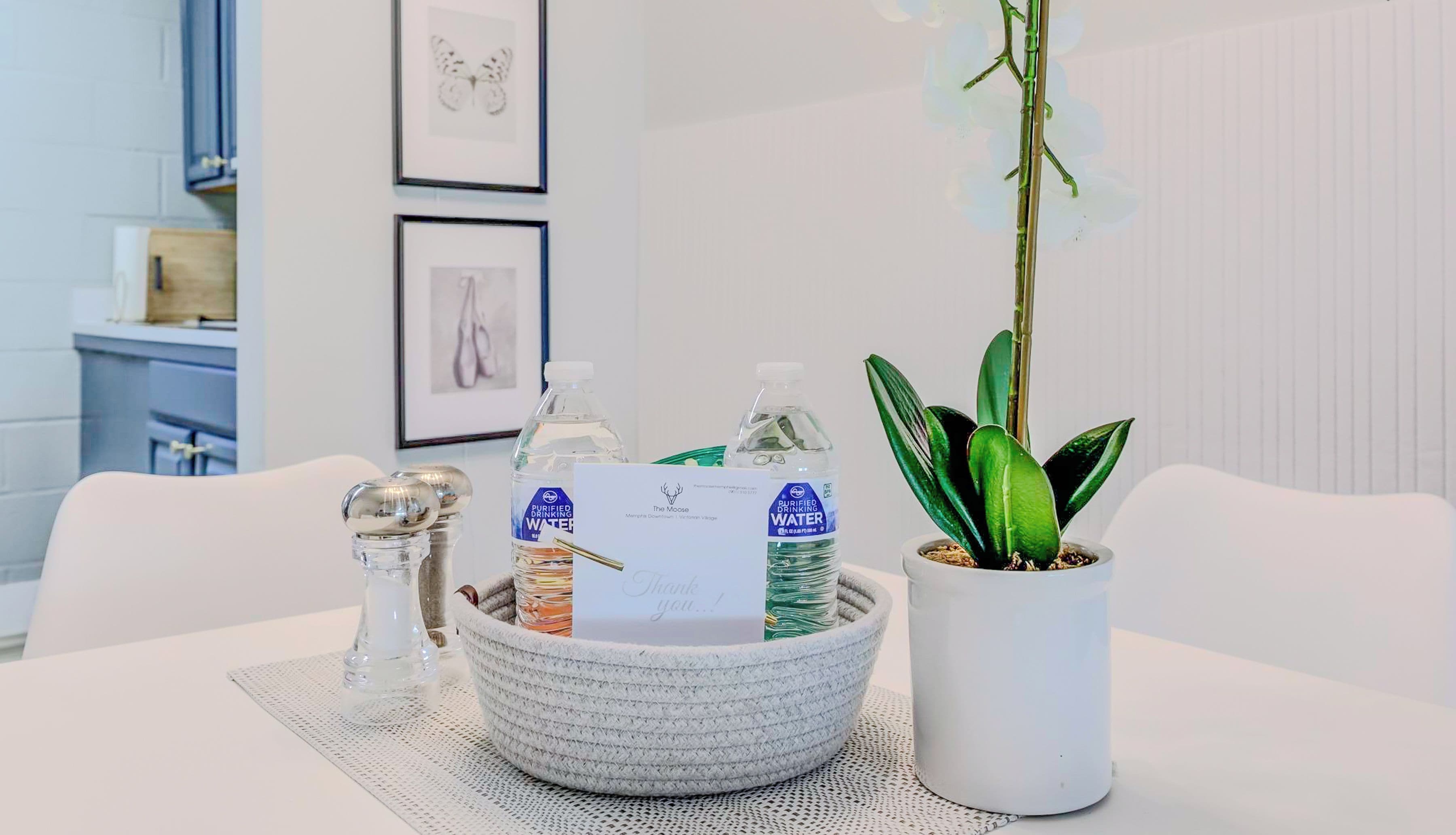 Une table à manger blanche, sur laquelle on trouve un set de table en fibres tressées, un salier en verre, un bol blanc contenant des bouteilles d’eau et une carte de remerciement, ainsi qu’une plante en pot.