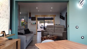 En moderne stue med en grå sofa, en beige lænestol og et træbord. Der er et spisebord i forgrunden, og man kan se udendørs gennem en glasdør.