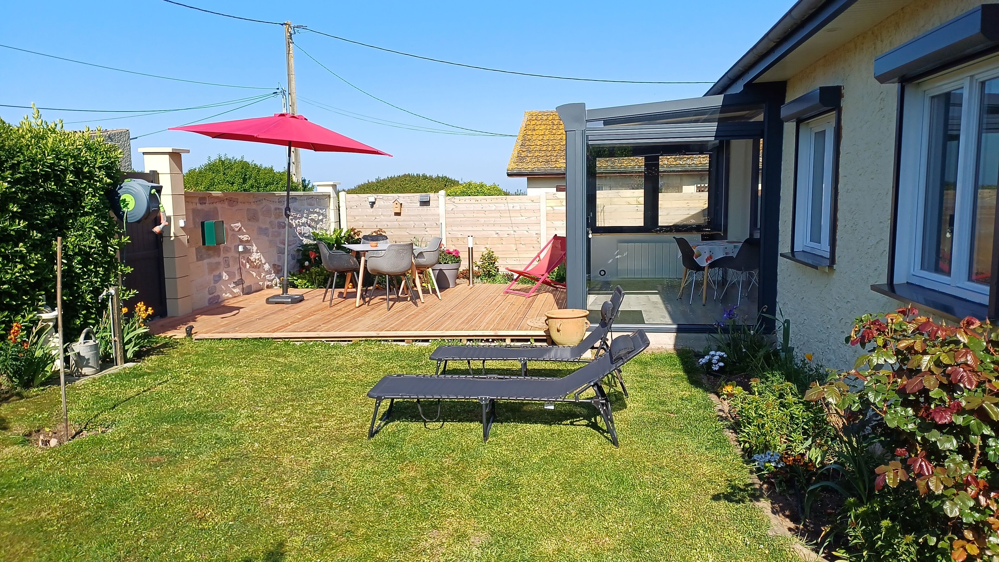 Un jardin avec une terrasse en bois, un parasol rouge et du mobilier d’extérieur.