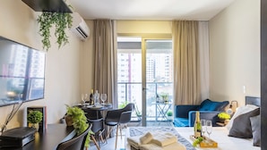A modern hotel room with a flat-screen TV, a dining area with a table and chairs, a bed with a view of the city, and a balcony with a table and chairs. - Allianz parque AC Solário | Brazilian Corner. (São Paulo)