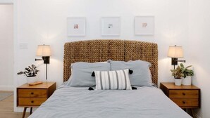 Een slaapkamer met een geweven hoofdbord, twee nachtkastjes met lampen, en drie ingelijste foto's aan de muur.