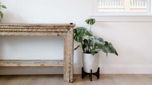 Een rustieke houten bank met een decoratieve rugleuning, een bloempot met grote bladeren en een raam met uitzicht op een balkon.
