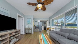 Un soggiorno con una grande TV a schermo piatto, una ventola a soffitto, un divano, una tavola da surf e vista sulla cucina e sala da pranzo.