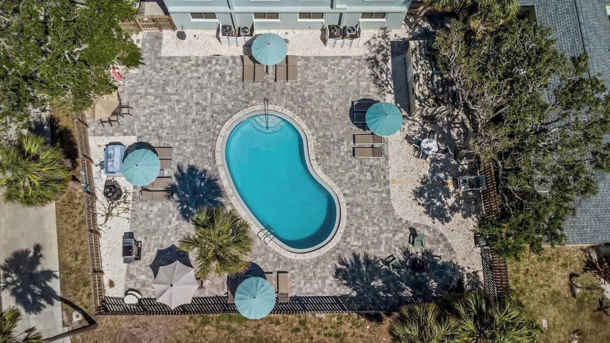 Vue aérienne d’un espace piscine avec des chaises longues et des parasols, entouré d’arbres et d’un bâtiment.