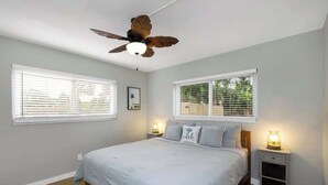 Une chambre à coucher comprenant un lit, deux tables de chevet avec des lampes, un ventilateur de plafond et deux fenêtres équipées de stores.