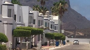 A coastal street with modern white buildings, palm trees, and a mountain in the background. - Casa Caracol, Apartamento Totalmente Equipado Para su Estancia en San Carlos Son (San Carlos Guaymas)