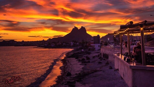 A coastal sunset with a mountain in the background, a beach, and a restaurant with outdoor seating. - Casa Caracol, Apartamento Totalmente Equipado Para su Estancia en San Carlos Son (San Carlos Guaymas)