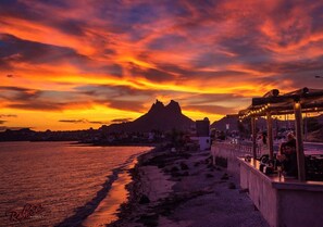 Een kustzonsondergang met een berg op de achtergrond, een strand en een restaurant met terras.