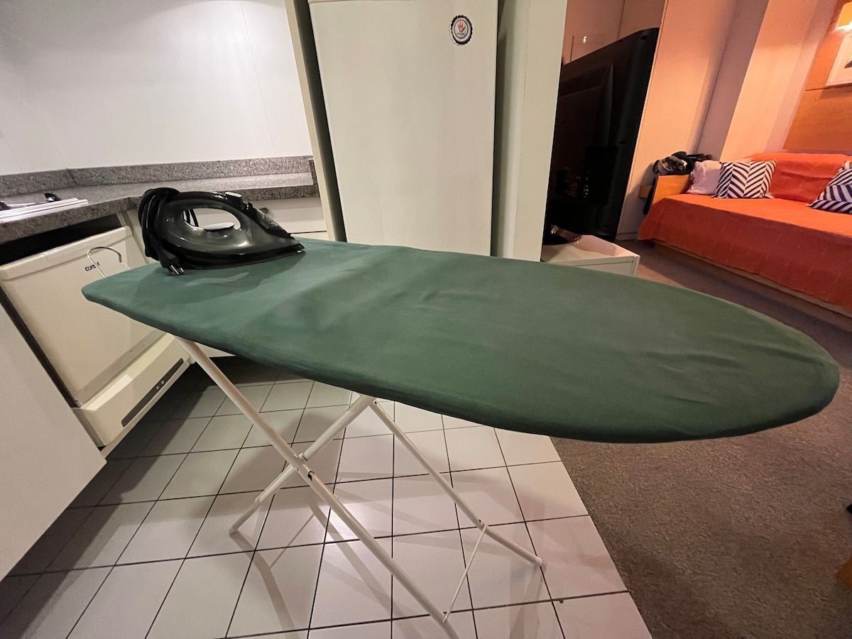 Een keuken met witte kasten, een groen strijkplank en een rode bank op de achtergrond.