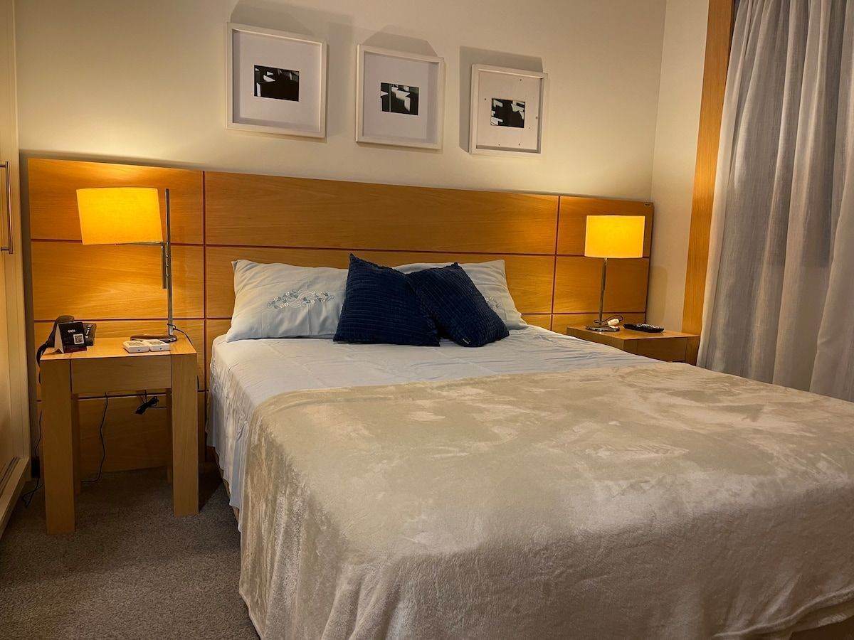 Quarto de hotel com uma cama grande, duas mesas de cabeceira, um criado-mudo e três quadros na parede.