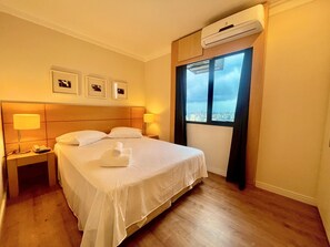 Habitación de hotel con cama, dos lámparas de noche, mesita de noche, unidad de aire acondicionado y ventana con vista a la ciudad.