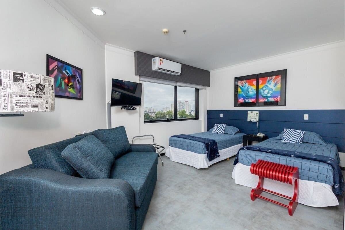 Habitación de hotel con un sofá gris, dos camas individuales con ropa de cama azul, un taburete rojo, un equipo de aire acondicionado, un televisor y vistas a la ciudad.