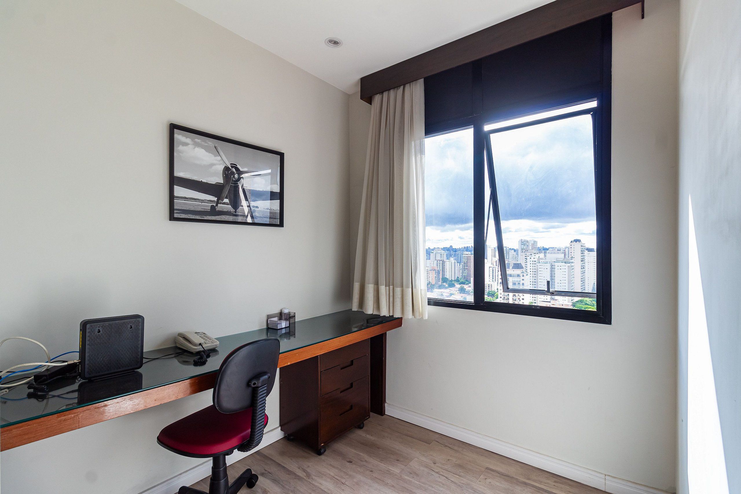 Un espace de bureau moderne avec un bureau en verre, un fauteuil de bureau rouge, un téléphone et un haut-parleur. Un tableau encadré est accroché au mur, et une fenêtre avec des rideaux offre une vue sur le paysage urbain.
