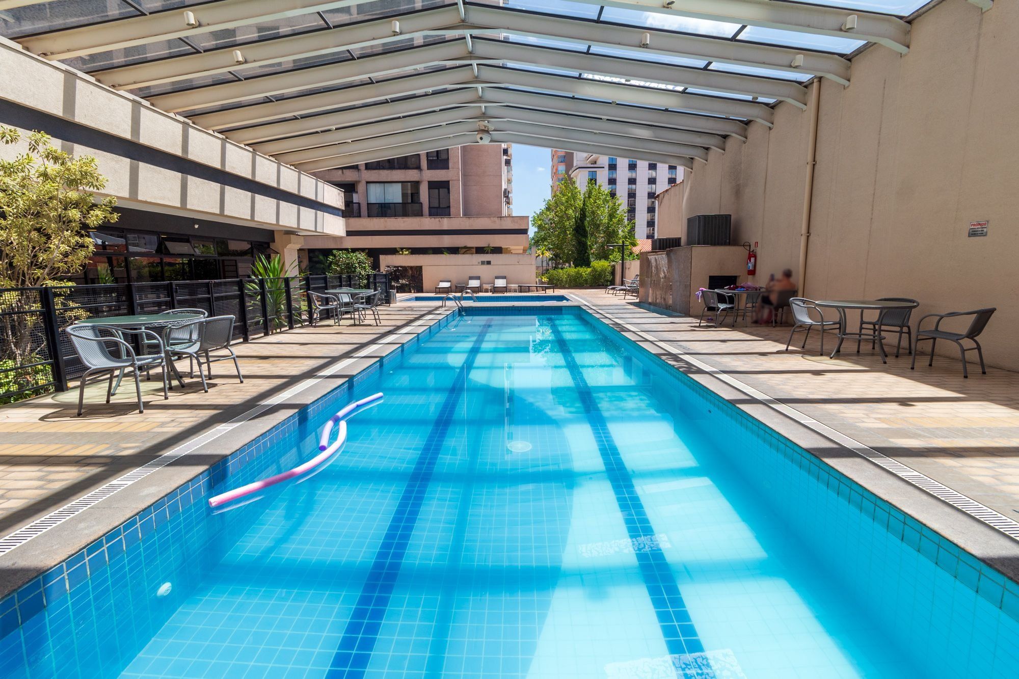 Uma piscina com teto de vidro, rodeada por espreguiçadeiras e mesas.