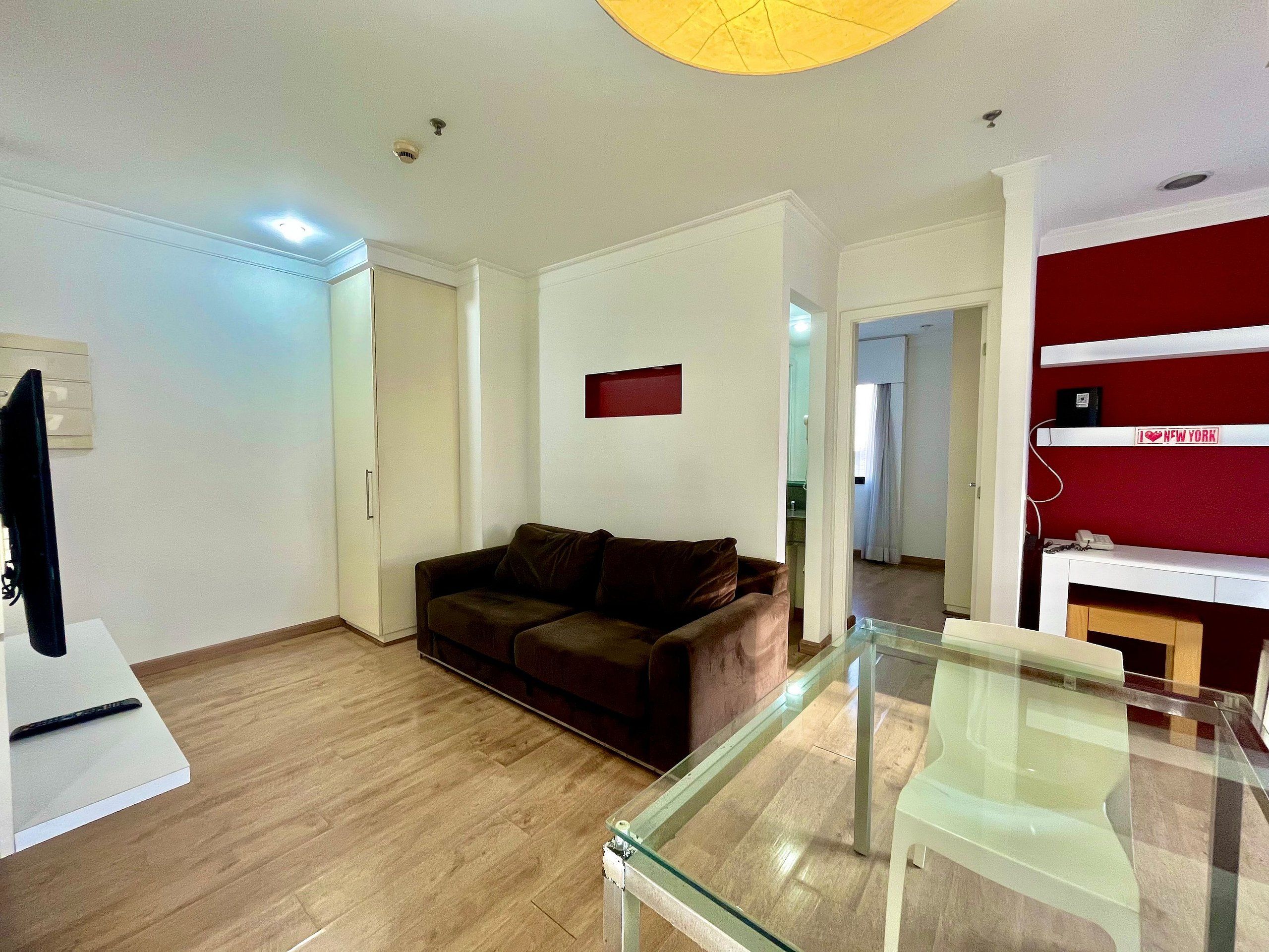 Uma sala de estar moderna com um sofá marrom, uma mesa de centro de vidro e uma TV sobre um móvel.