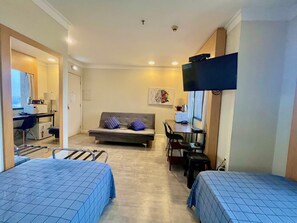 Une chambre d’hôtel compacte comprenant deux lits simples, un canapé, un bureau avec une chaise, une télévision et une petite kitchenette.