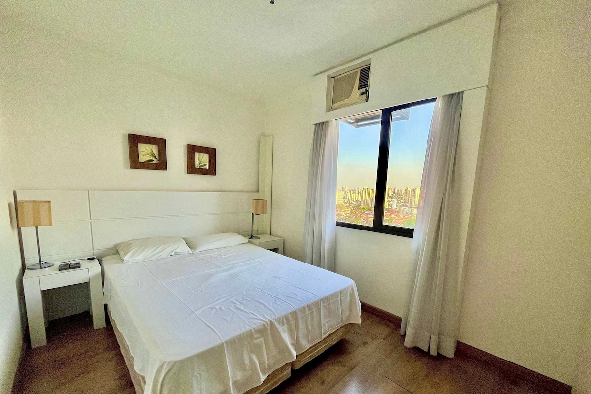 Una habitación de hotel con una cama grande, dos mesitas de noche con lámparas, una ventana con cortinas y vistas a la ciudad.