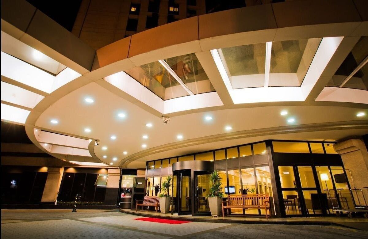 A modern hotel entrance with a curved glass facade, a red carpet, and a central seating area.
