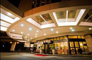 Une entrée d’hôtel moderne avec une façade en verre incurvé, un tapis rouge et un espace salon central.