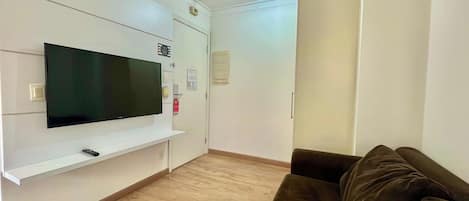 Una sala de estar con una televisión de pantalla plana montada en la pared, un control remoto sobre un pedestal, un sofá marrón y piso de madera.