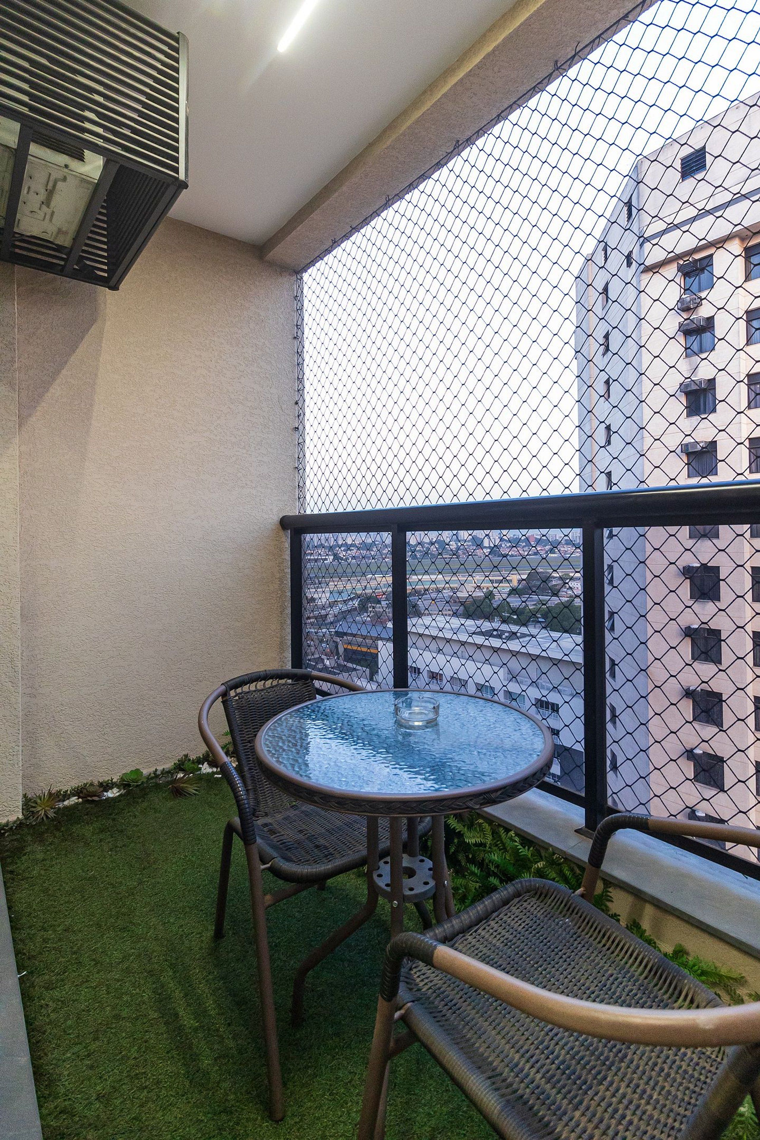 Varanda com mesa de vidro e cadeiras de vime, grade de tela e vista para a cidade.