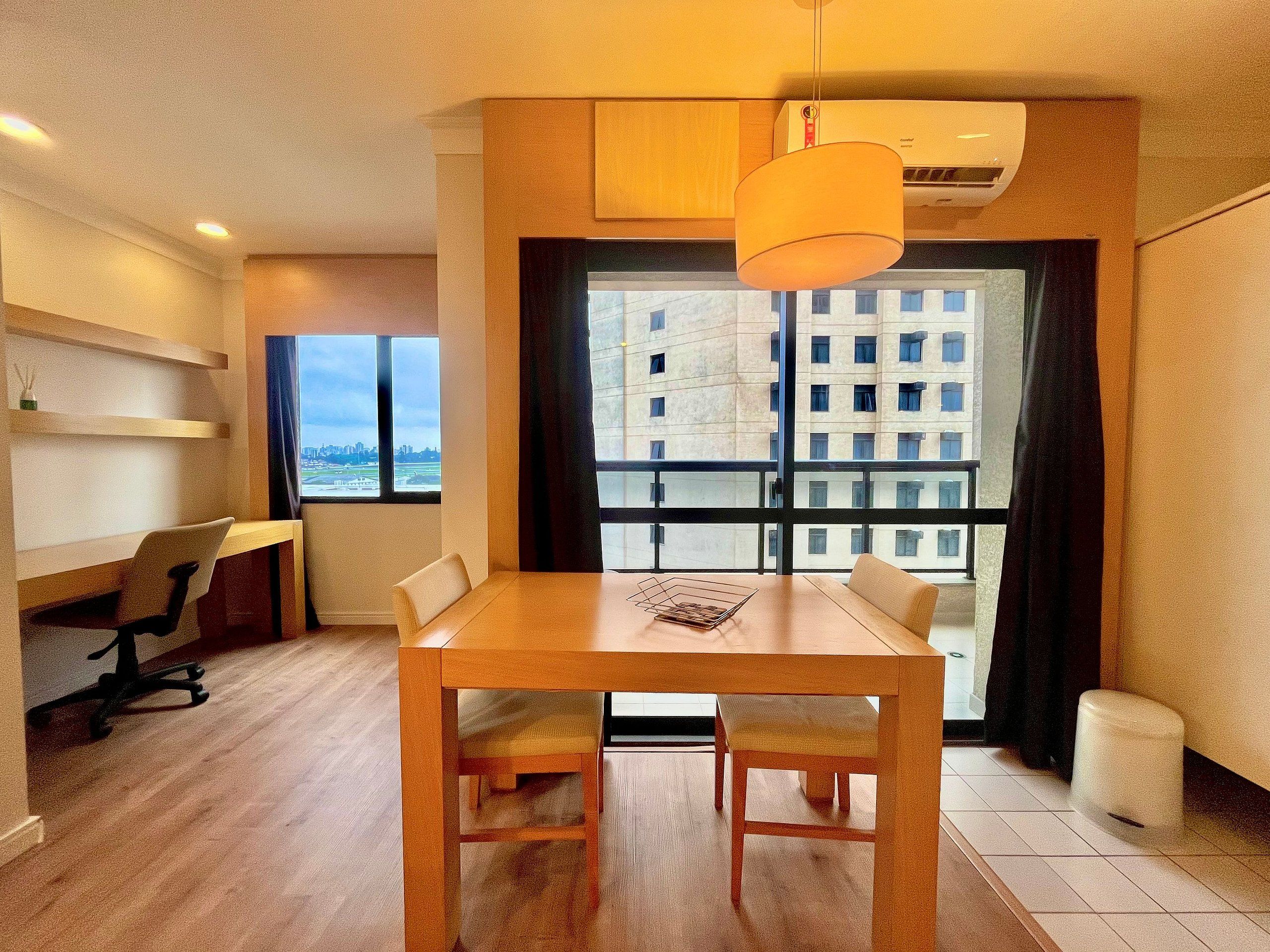 A modern dining area with a wooden table and chairs, a hanging light fixture, and a view of a cityscape through large windows.