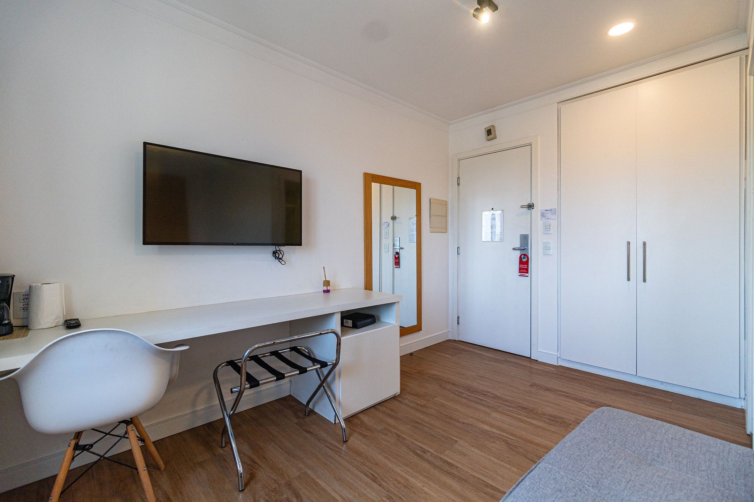 Quarto moderno com televisão de tela plana, escrivaninha branca, cadeira branca, piso de madeira e guarda-roupa.