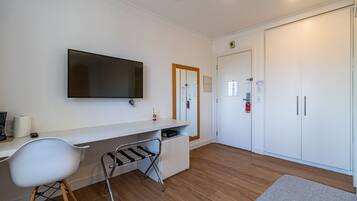 Quarto moderno com televisão de tela plana, escrivaninha branca, cadeira branca, piso de madeira e guarda-roupa.
