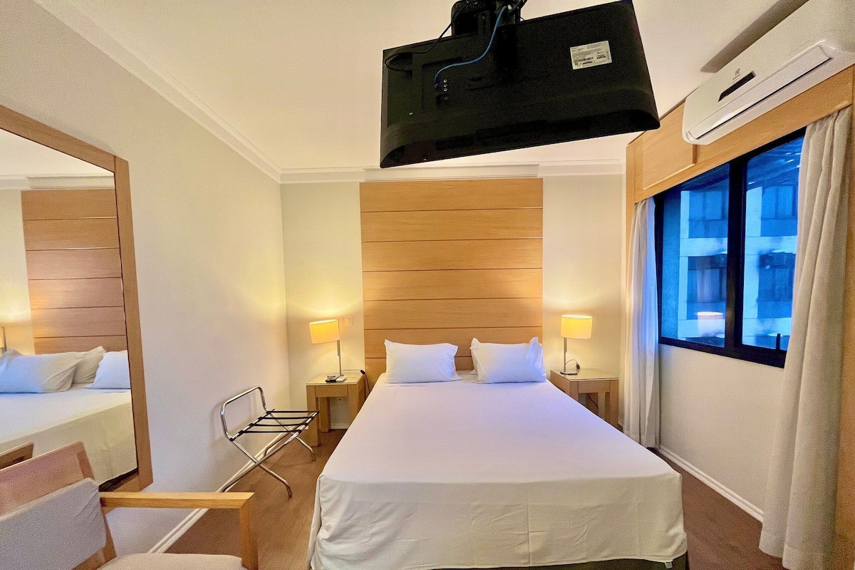 Habitación de hotel con una cama grande, dos mesitas de noche con lámparas, una silla, un espejo y una ventana con cortinas.