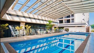 A modern indoor swimming pool with clear blue water, surrounded by white lounge chairs and a glass balustrade.