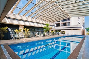 Una piscina interior moderna con agua azul cristalina, rodeada de sillones de descanso blancos y una barandilla de vidrio.