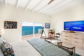 A bedroom with a bed, a TV, a desk, and a chair. There is a balcony with a view of the ocean.
